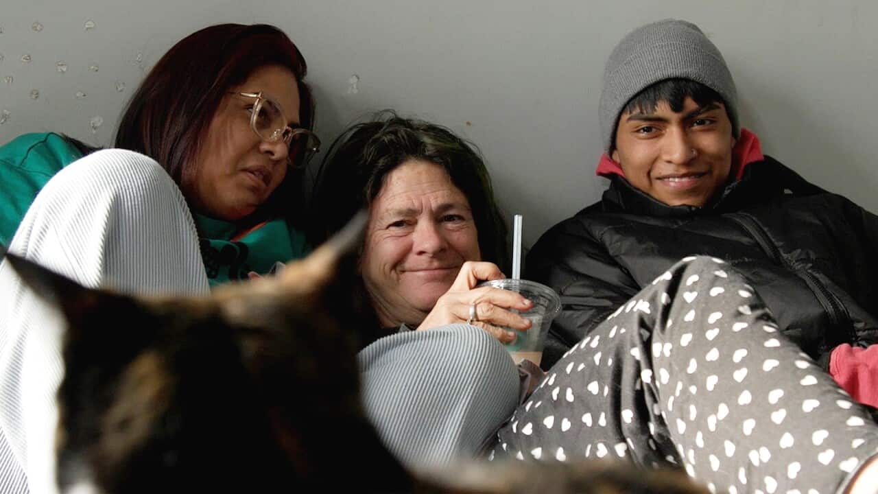 A woman sitting on a bed, holding a plastic cup with a straw, and smiling with a young woman and a man by her side.
