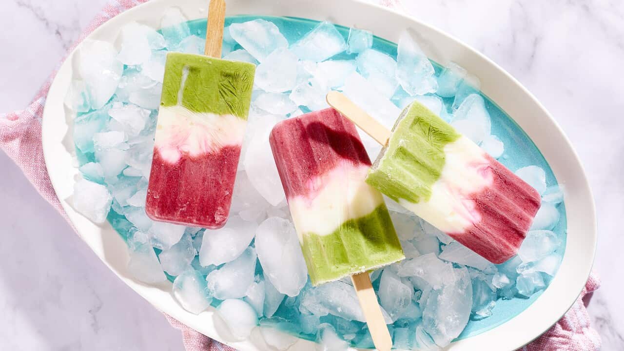 A blue platter with a white rim is filled with chunks of ice. Three popsicles sit on the ice; each has three layers, green, white and red. The tray sits in a striped pink cloth napkin on a pale marble surface.