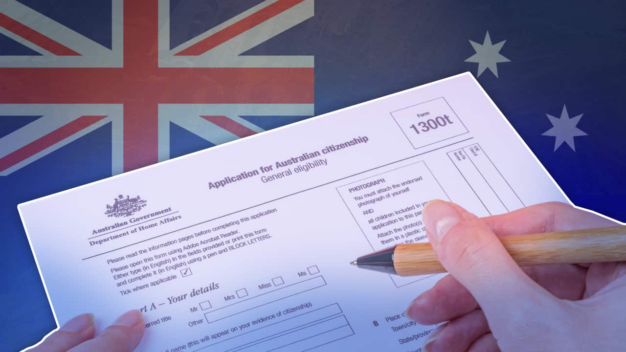A hand holding a pen hovers above an Australian citizenship application form, against the backdrop of an Australian flag.