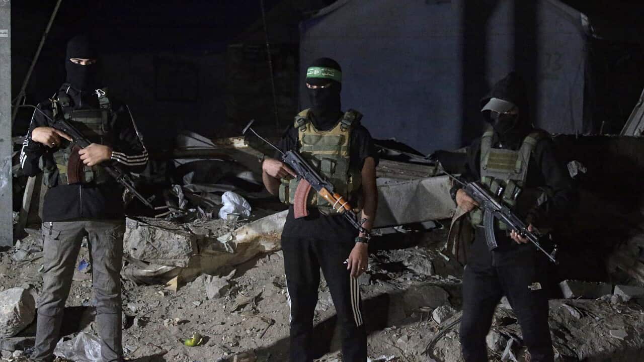Three armed men wearing face coverings and tactical vests stand at night amidst rubble, holding what appear to be AK-style assault rifles. They are positioned in front of a partially visible damaged building.