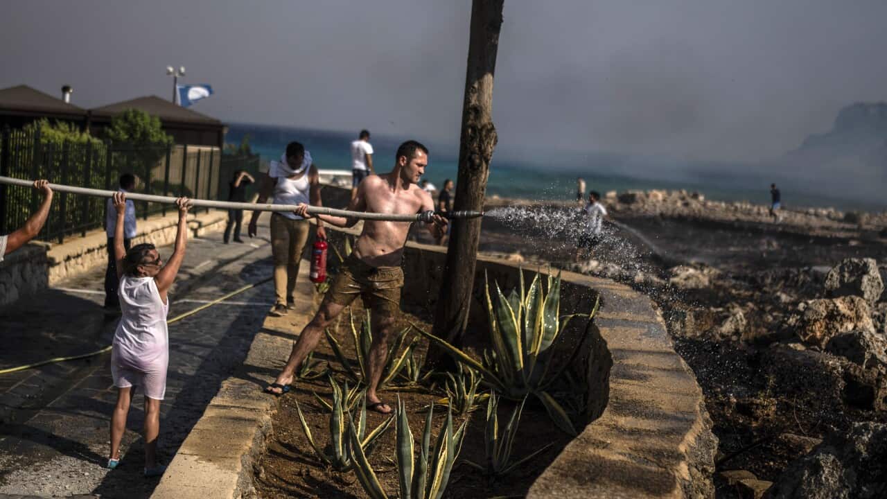 A man in shorts and a woman with wet clothes hold a water hose, with a smoky background behind them.