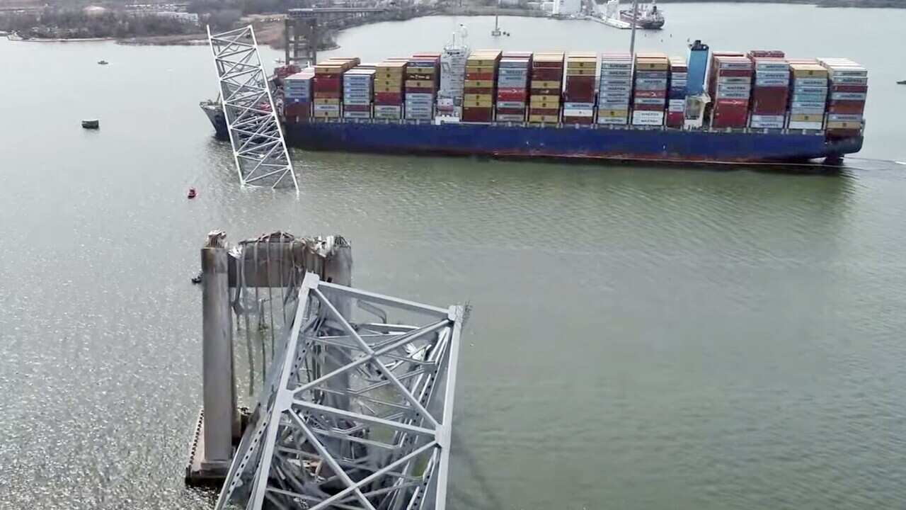 A container ship with collapsed sections of a metal bridge laying over the bow. Other fragments of the bridge sit askance alongside a broken pylon.