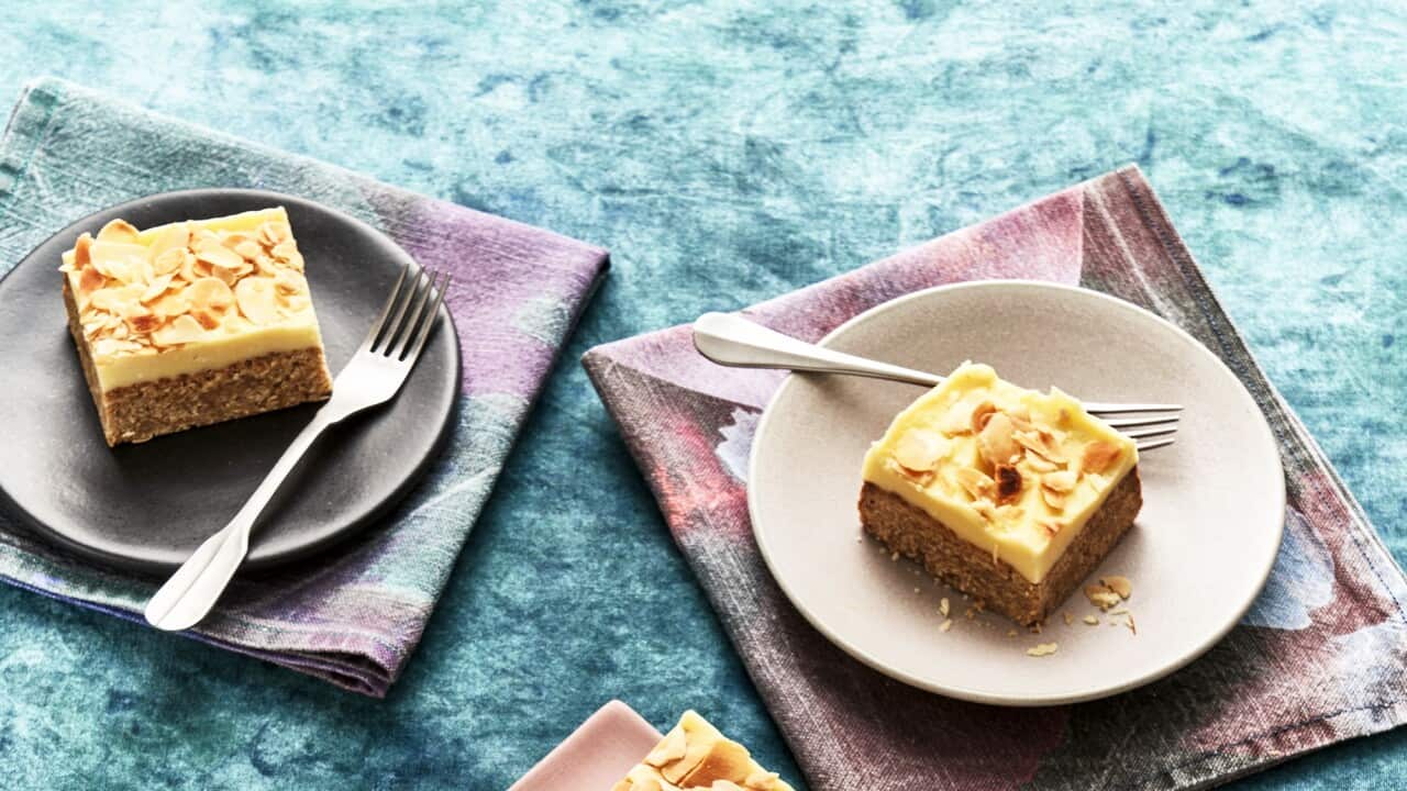 Deep squarish pieces of a layered bake, with a thick brownish bottom later and creamy top layer, scattered with flaked almonds, sit on wide plates. Each plate sits on a napkin in tones of blue, grey and pink.