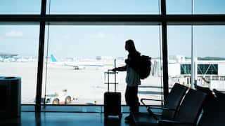 A woman with a suitcase standing in front of a large window at an airport, with planes and the tarmac visible behind her.