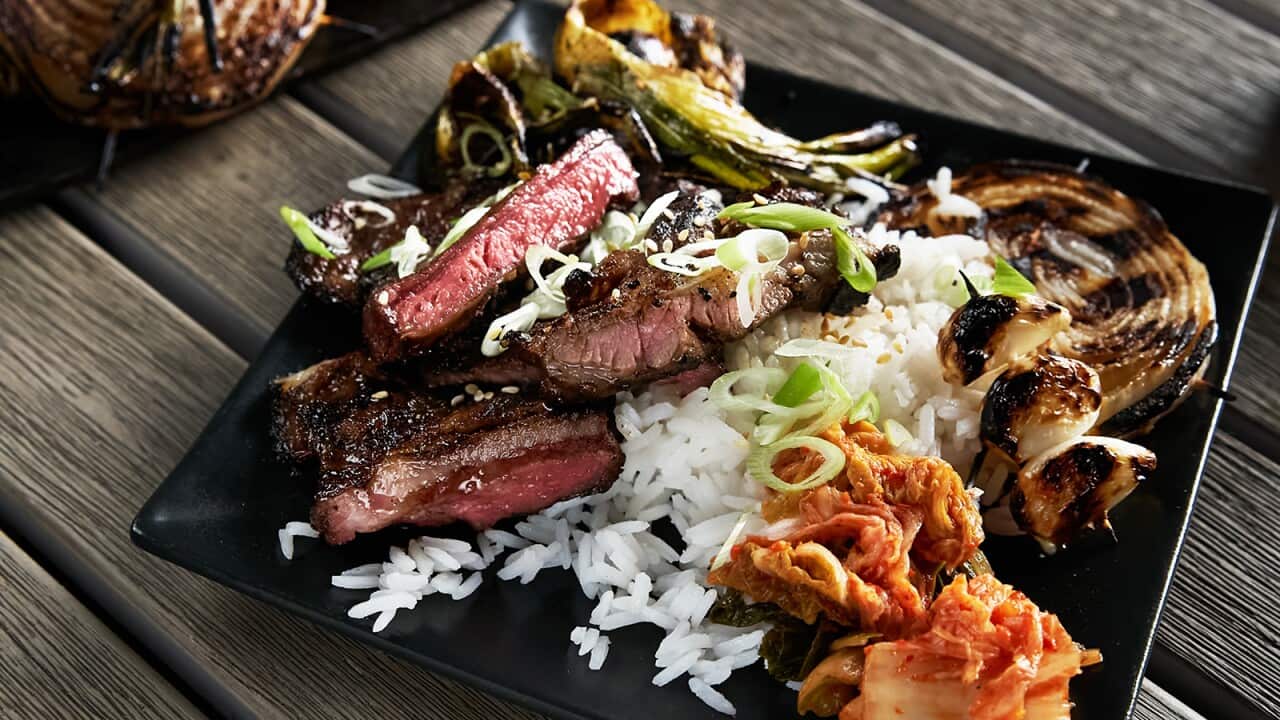 White rice, cooked beef, kim chi and charry onion and green onion sit on a square black plate, on a wooden surface.