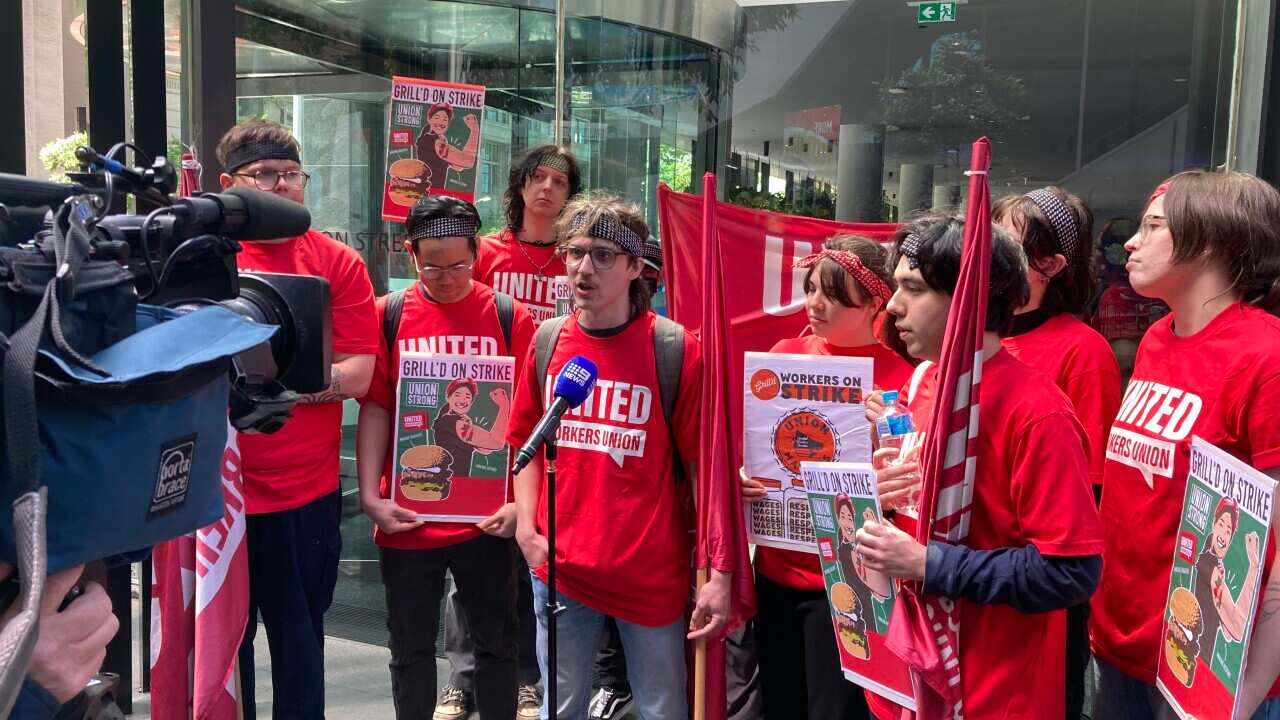Several young people wearing red T-shirts with United Workers Union emblazoned on the front stand behind a microphone. Some are carrying placards with phrases like “Grill'd on strike“ and “workers on strike“
