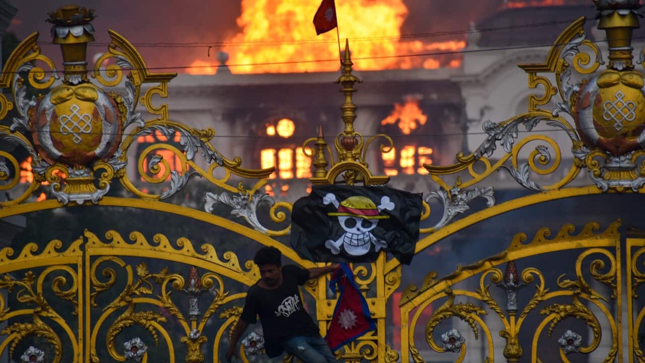A large building is engulfed in flames behind a golden gate, with a man climbing the gate's bars. He is holding a Nepali flag and a pirate flag featuring a skull with a straw hat is hanging from the gate.