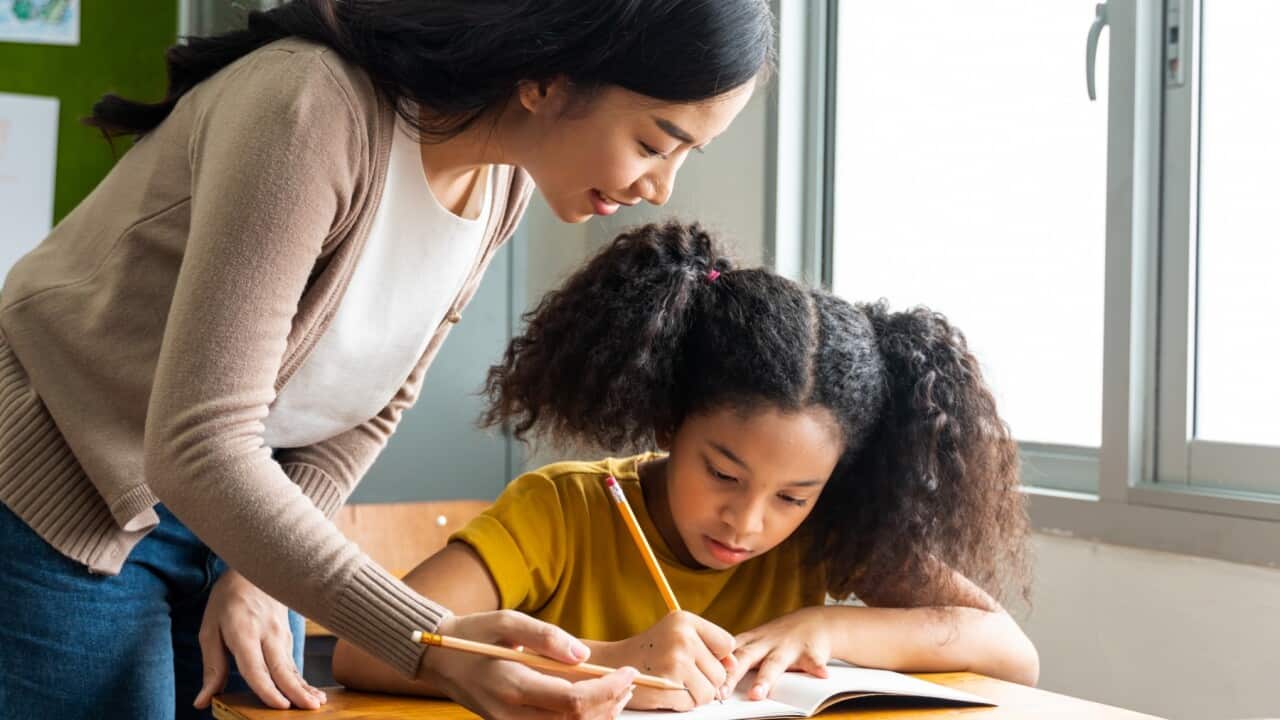 Asian school teacher assisting female student in classroom. Young woman working in school helping girl with her writing, education, support, care -