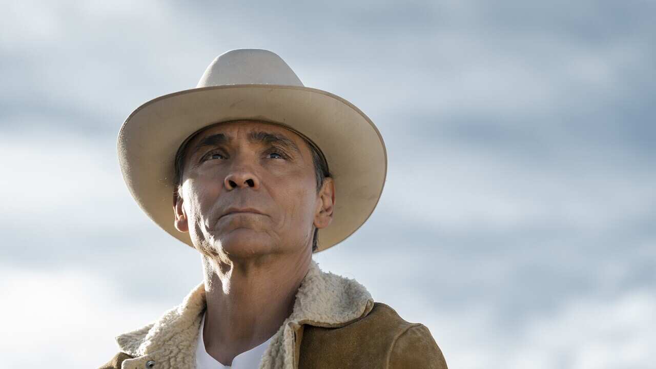 Seen slightly from below, with head and shoulders visible, actor Zahn McClarnon stands looking pensively ahead. The out of focus blue-white background adds a sense of occasion.