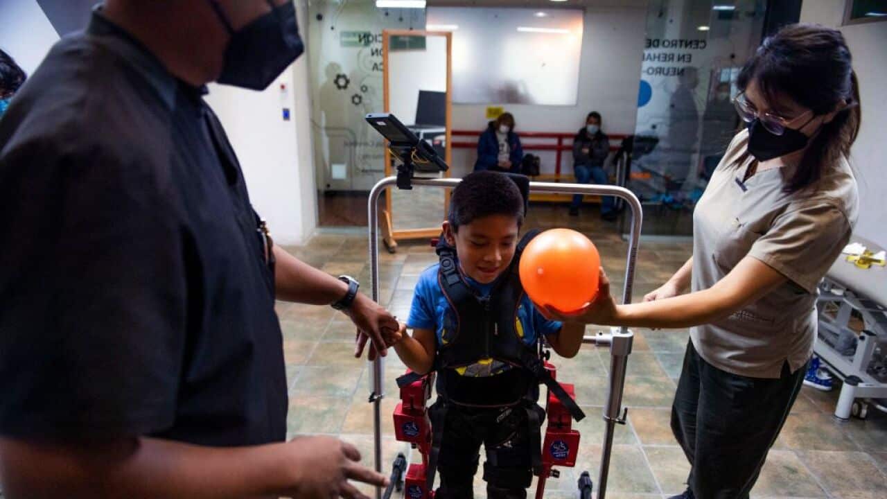 An eight-year-old boy with cerebral palsy in a robotic frame that helps him to walk. Two adults are standing nearby