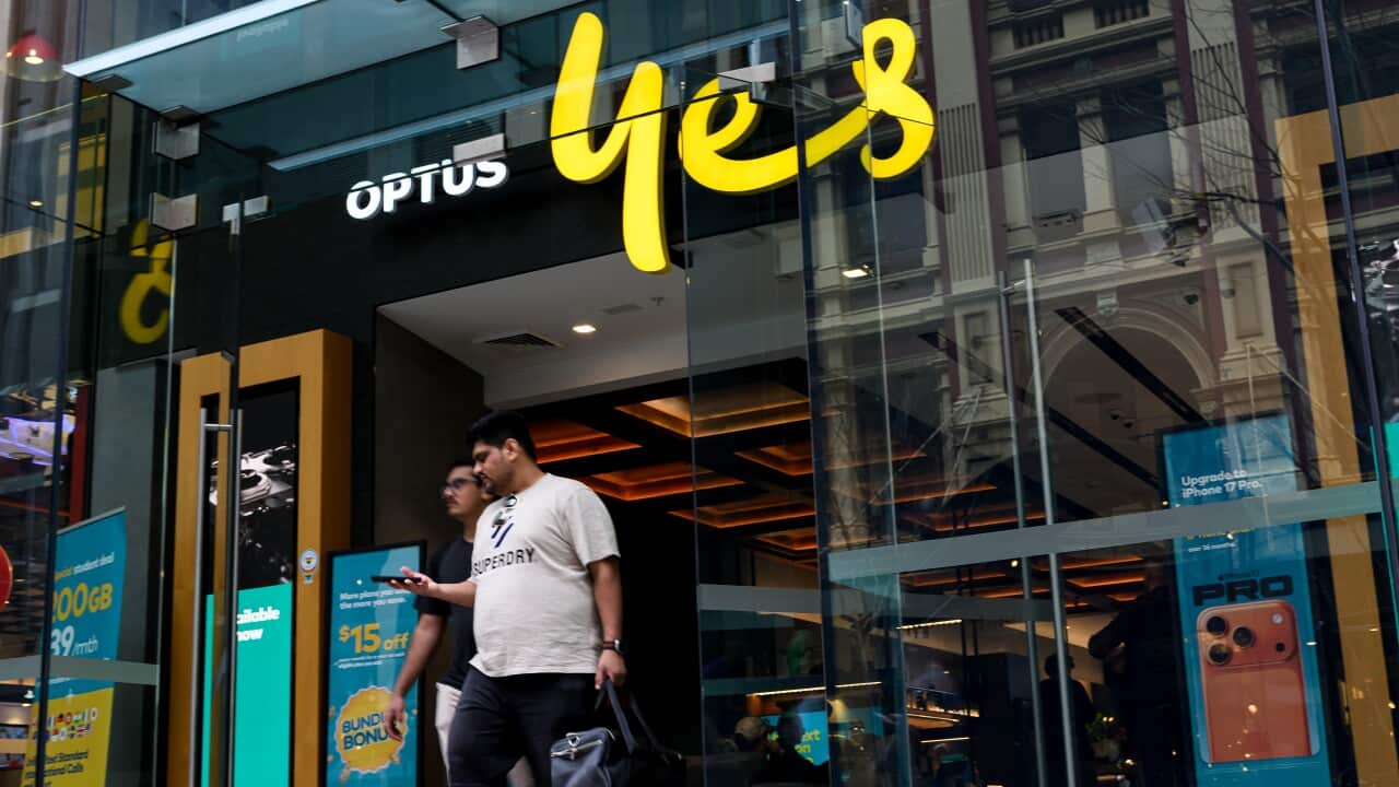 A wide-angle street-level shot shows the entrance to an Optus store with the company's "yes" logo in bright yellow above the door, as two men walk out of the glass entrance.