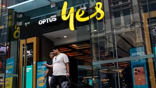 A wide-angle street-level shot shows the entrance to an Optus store with the company's "yes" logo in bright yellow above the door, as two men walk out of the glass entrance.