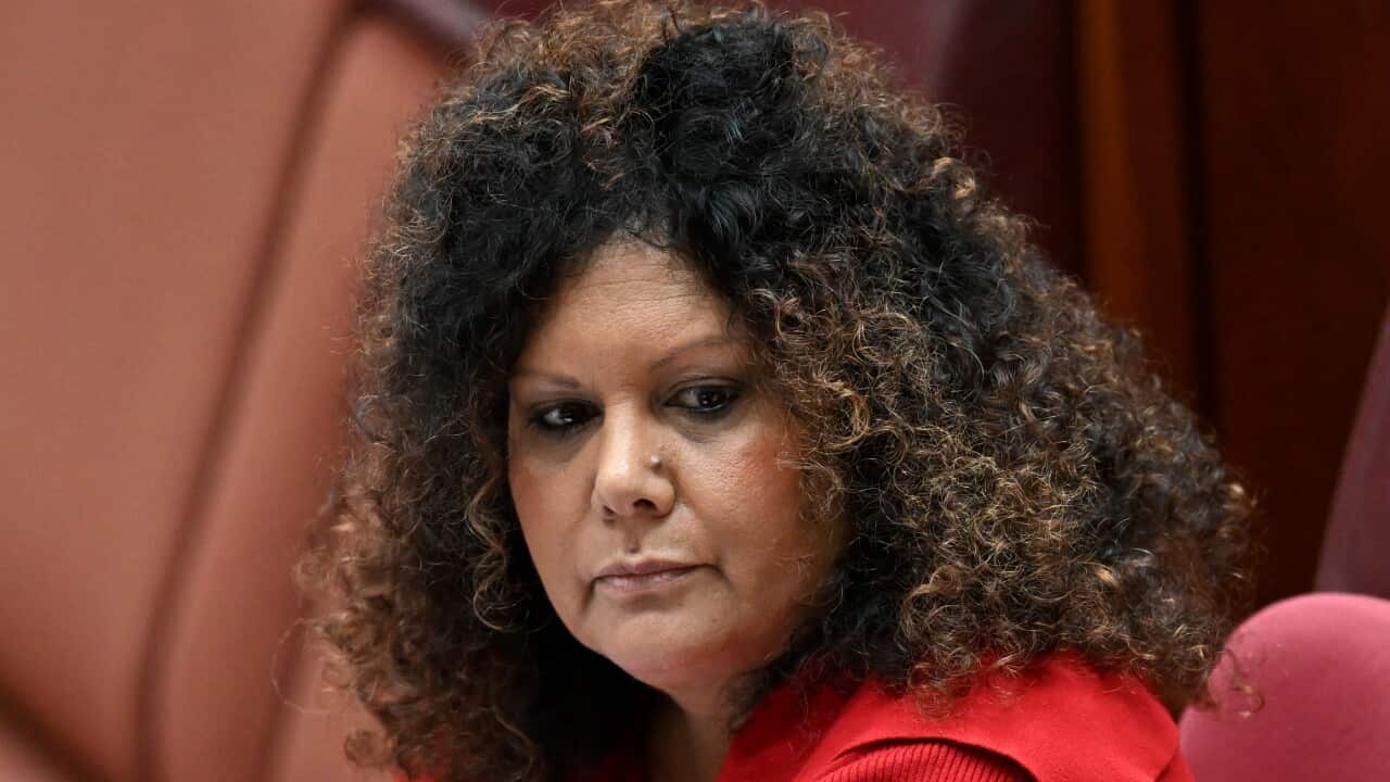 A woman in a red jumper with dark curly hair looks to her left with a solemn expression.