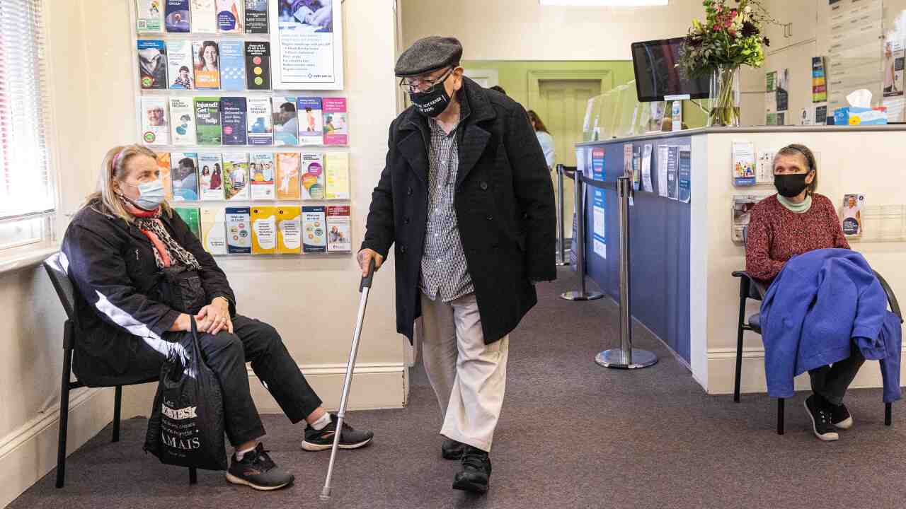 Two woman sitting at a doctor's surgery and a man walking through with a walking stick