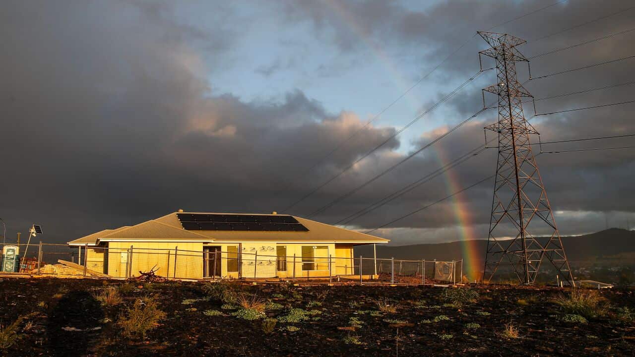 A rainbow arches over a newly built, single-story home with solar panels on its roof. The house is on a hill surrounded by scorched earth, with power lines and a large transmission tower in the foreground.