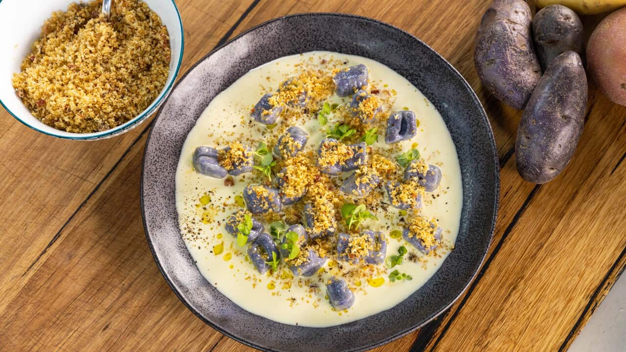 A wide silver-brown pottery bowl sits on a wooden surface. It holds purple gnocchi in a creamy sauce. A bowl of cooked breadcrumb topping sits alongside. Some has been sprinkled on the gnocchi, along with small herb leaves.