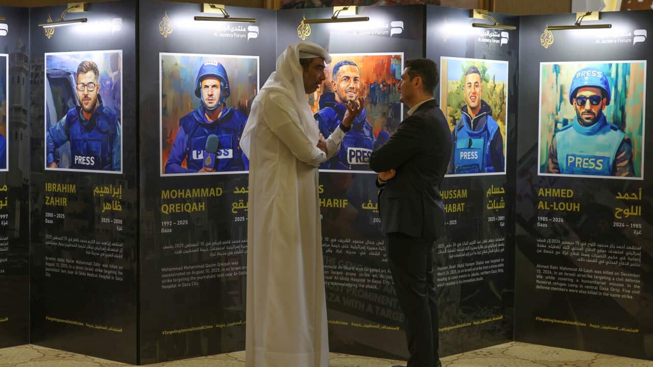 Two men speaking in front of a display showing images and biographies of five men in press jackets