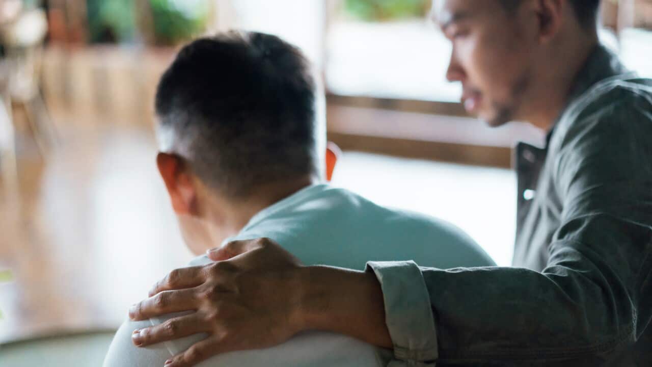 Rear view of son and elderly father sitting together at home. Son caring for his father, putting hand on his shoulder, comforting and consoling him. Family love, bonding, care and confidence