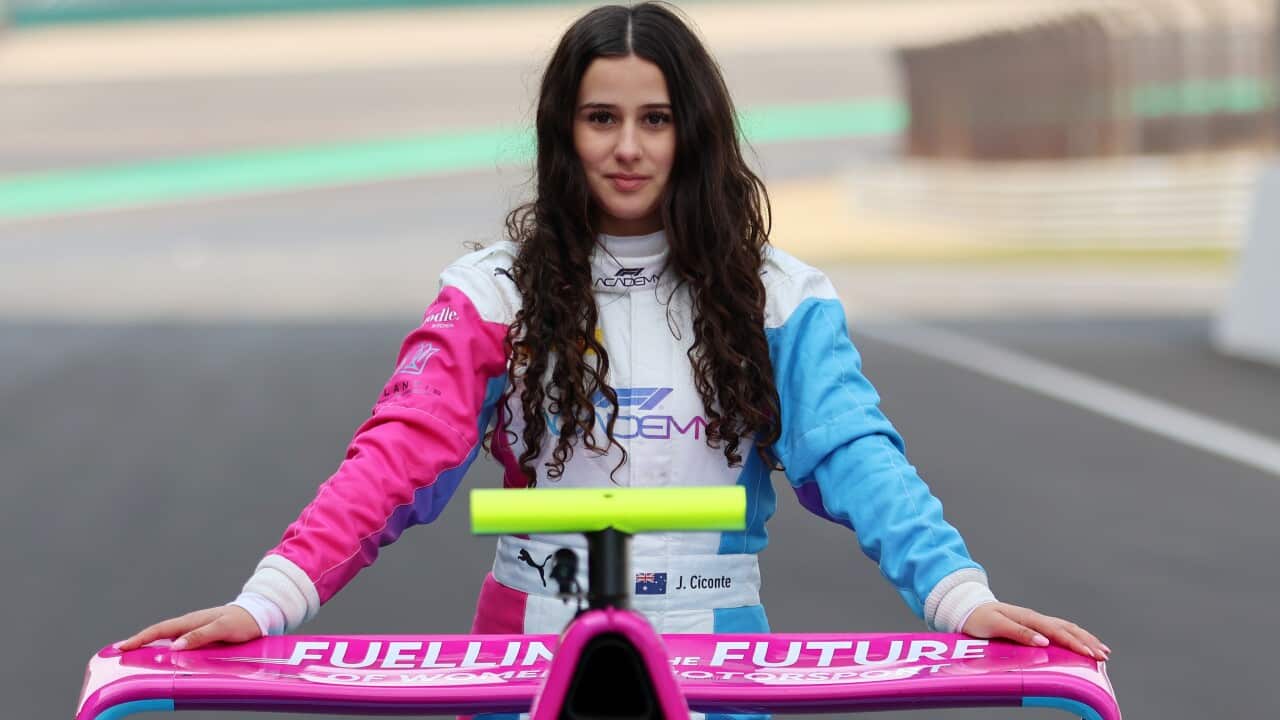 A teenage girl with long dark hair and wearing a white racing suit with one pink sleeve and one light blue sleeve stands on a race track behind the pink rear wing of an open-wheel racing car.
