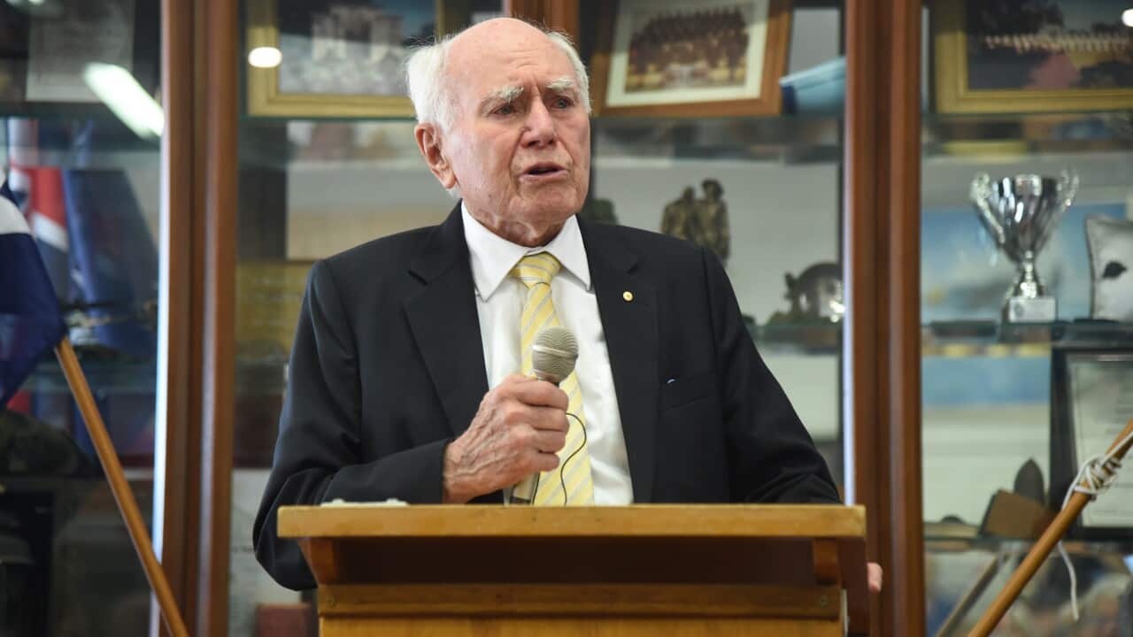 Former prime minister John Howard, stands at a wooden lectern, holding a microphone in his hand as he speaks.