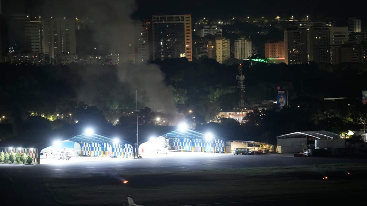 A high-angle nighttime view of an airfield featuring illuminated hangars and a small private jet, with dark smoke rising from behind the hangars against a backdrop of city buildings.