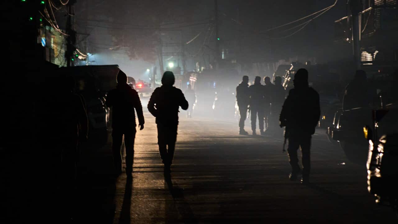 Silhouetted figures, including two walking toward the foreground and a group standing further back, are shrouded in thick fog or smoke on a dark street at night.