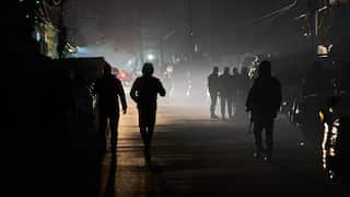 Silhouetted figures, including two walking toward the foreground and a group standing further back, are shrouded in thick fog or smoke on a dark street at night.