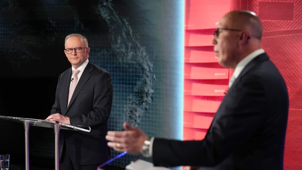Two men in suits stand at their own lecterns in a news studio. The man on the right is speaking while the other looks at him.