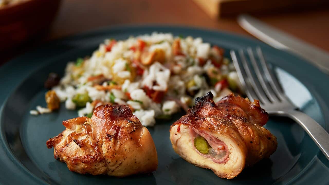 A cooked, stuffed chicken breast, cut in half, sits on a dark plate, with a fork alongside. A mound of rice, studded with small pieces of variously coloured vegetables, sits behind the chicken.