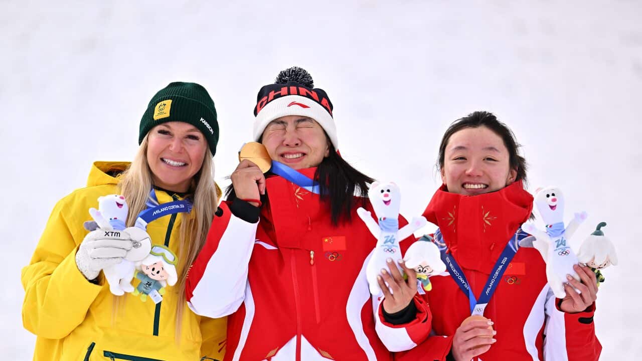 Three female Olympic medalists in winter gear smile on a podium, holding their medals and mascots.