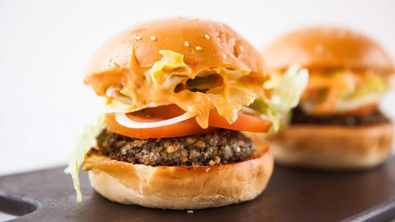 A brown patty with tomato, lettuce, onion rings and sauce, sits in a bun. Another can be seen, out of focus, behind it.