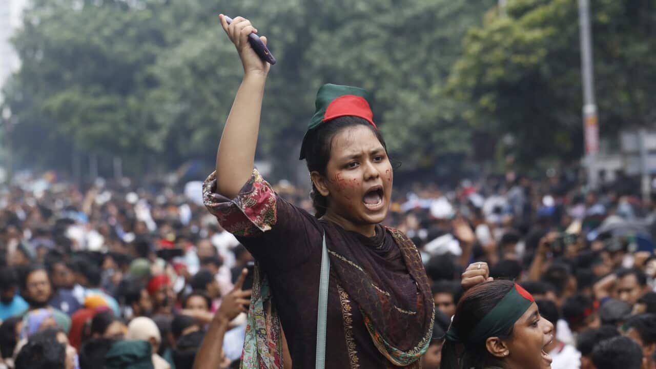 A woman wearing a brown sari raises a hand clasping a mobile phone in the air. Her mouth is open as if shouting and she is surrounded by a large crowd.