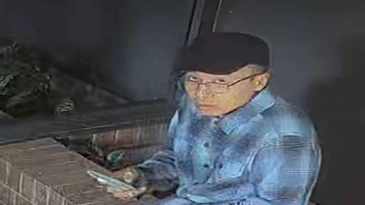 A CCTV grab of an Asian man wearing glasses, a black hat and blue shirt holding a phone