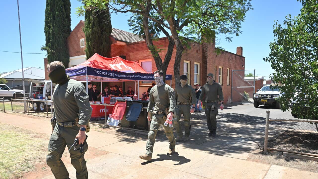 Four men in olive-green tactical gear and face gaiters walk along a paved path in front of a red brick building and a Rapid Relief Team tent.