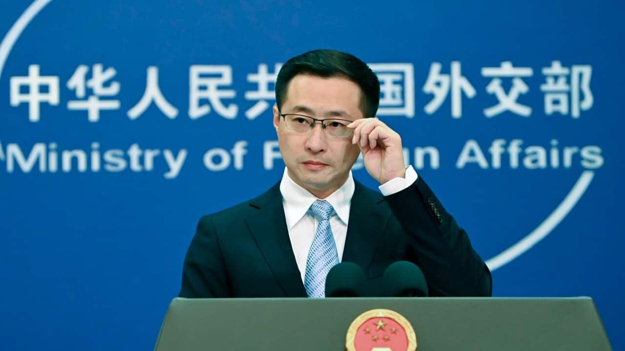 A man in a suit and glasses stands at a podium featuring the national emblem of China, speaking in front of a blue backdrop for the Ministry of Foreign Affairs.