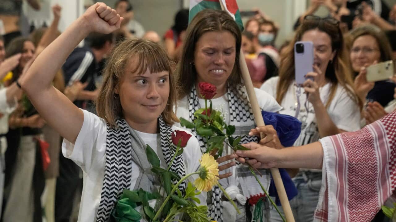 A woman in a white T shirt and scarf and holding flowers holds her hand up, seemingly in victory, while surrounded by crowds of others.