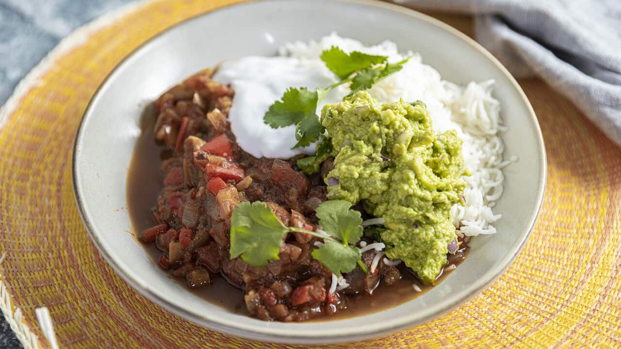 A pale bowl sits on a vibrant yellow circular mat. The bowl holds a vegetable stew, rice, gaucamole and cream, garnished with coriander leaves.