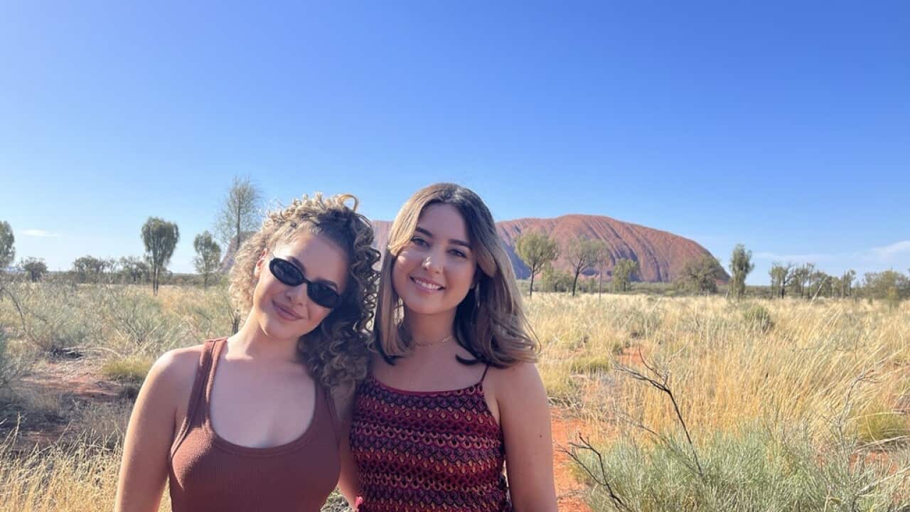 Two smiling young women stand in the central australian desert, with uluru in the background