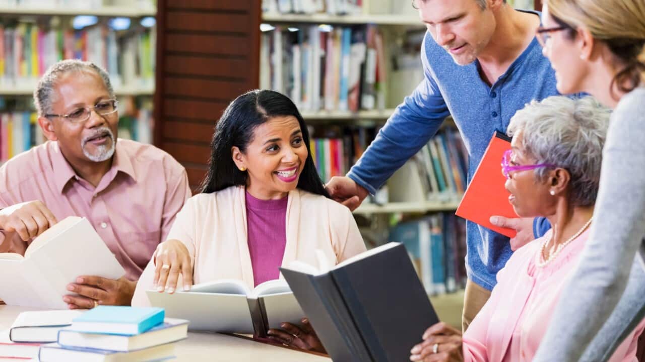 A diverse group of adults taking a continuing education class, sitting in a library looking at books.