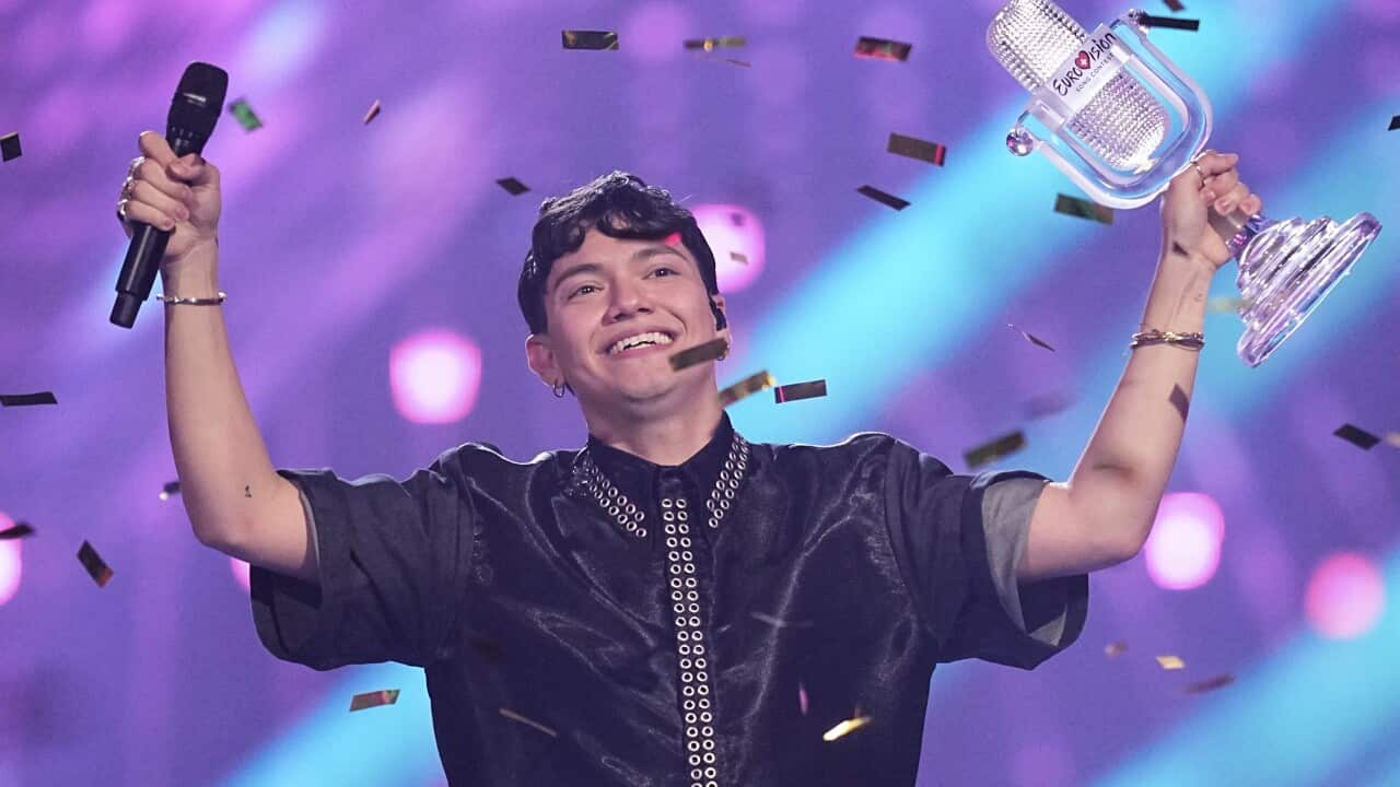 A person celebrates on stage holding a Eurovision trophy and a microphone, surrounded by confetti.