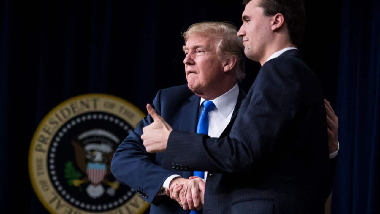 An older man with blonde hair wearing a navy suit, shaking hands with a younger man giving a thumbs up.