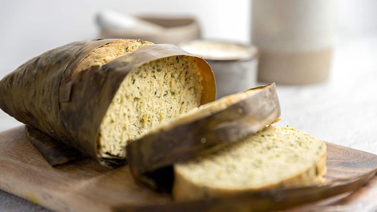 A damper, wrapped in the banan leaf it was cooked in, sits on a wooden board. Several slices have been cut, showing the green-flecked golden interior.