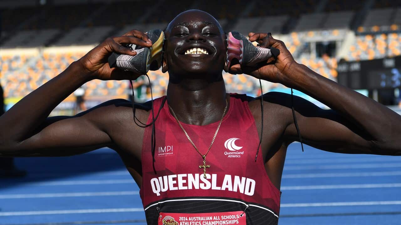 A young athlete wearing a red jersey with "Queensland" holds his running shoes up to his ears. He has a wide smile on his face.