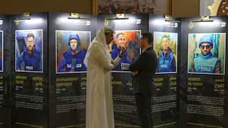 Two men speaking in front of a display showing images and biographies of five men in press jackets