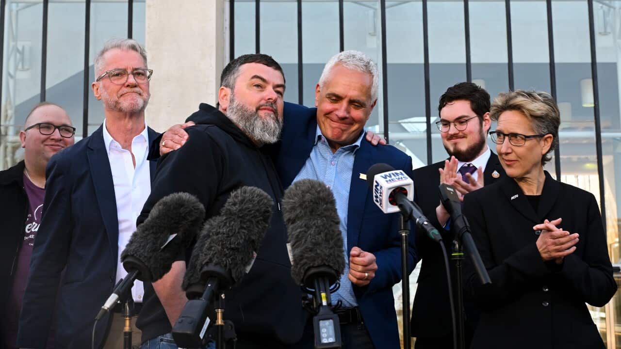 A group of people speak to the media as a man in the middle smiles and hugs another.