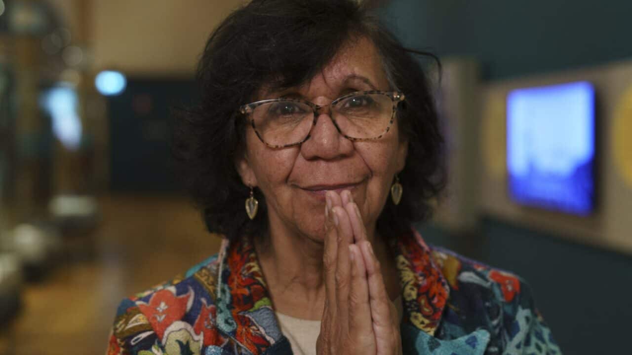 Nanny Nellie, an older Aboriginal woman with glasses and shoulder-length dark hair, stands indoors with her hands gently pressed together in front of her face. She wears a colorful patterned jacket and heart-shaped earrings, with a warm and thoughtful expression.