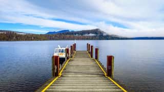 A small jetty extends into a lake, with a boat tied up on its side. In the distance, low clouds engulf wooded hills.