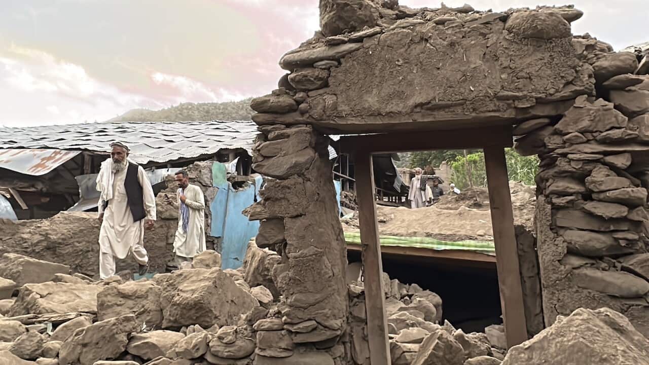 People walking past badly damaged mud-and-stone buildings.
