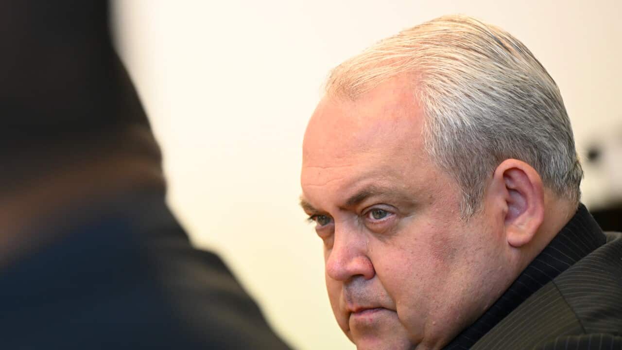 A close-up photograph of a middle-aged man with light eyes and slicked-back gray hair, wearing a pinstripe suit and looking intensely toward the foreground.