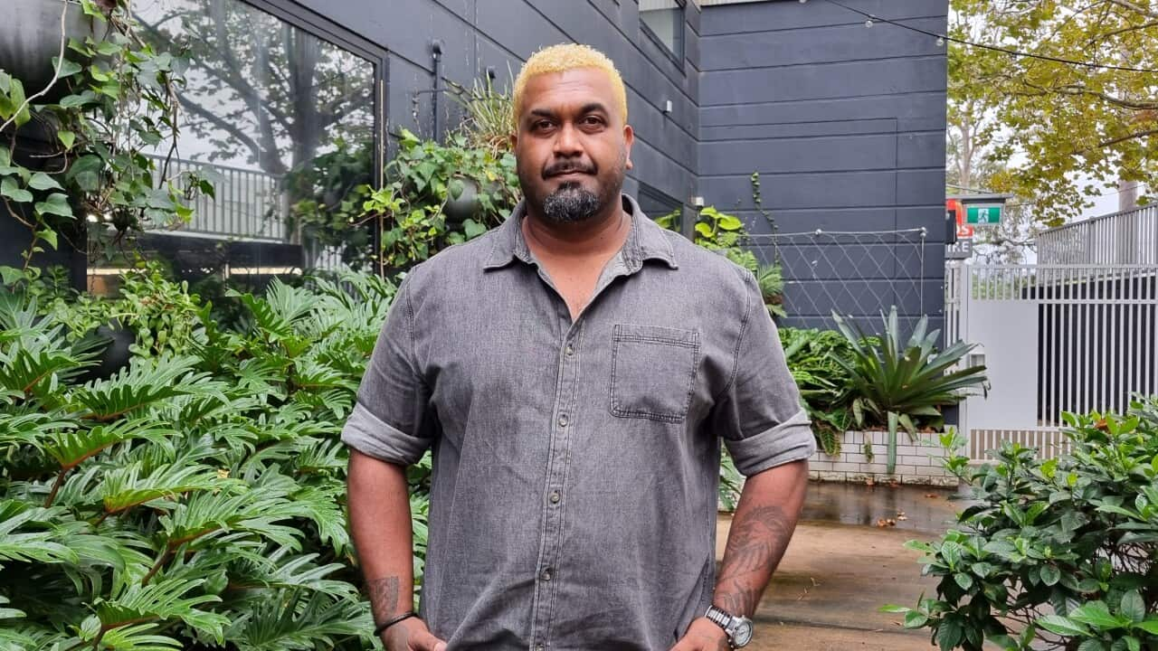 A man in a courtyard stands with his hands in his pockets, looking at the camera with a neutral expression.